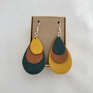 Tri Color Rainbow Earrings in Teal Brown and Yellow. This pair of earrings features a mix-matched order of color, so one earring is yellow, brown, and teal, whereas the other is teal, brown, and yellow. The earrings are pictured on a brown card in a brown jewelry box, which both come included with the earrings. They are pictured against a white background.