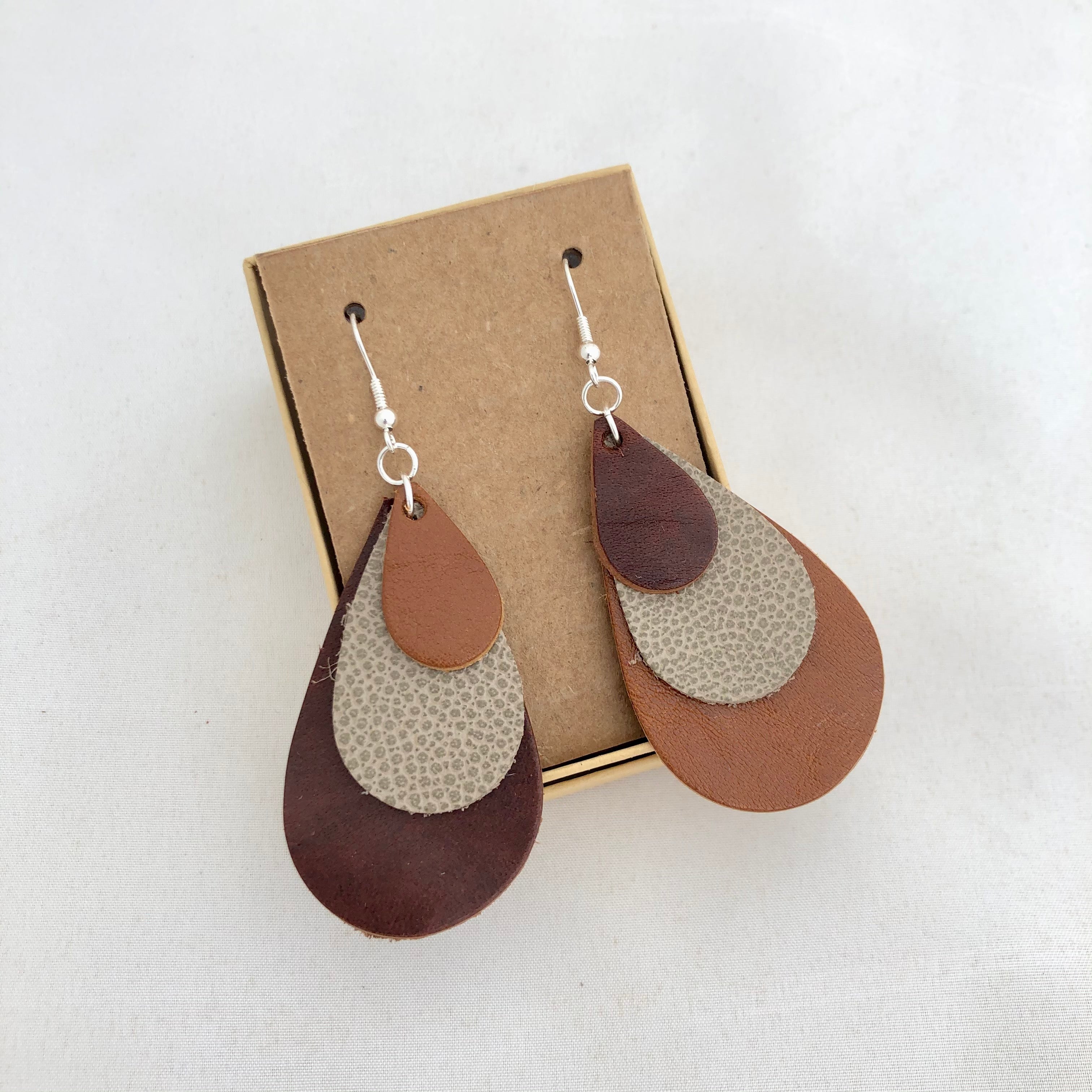 Tri Color Rainbow Earrings in Shades of Brown. This pair of earrings features a mix-matched order of color, so one earring is light brown, spotted tan, and dark brown, whereas the other is dark brown, spotted brown, and light brown. The earrings are pictured on a brown card in a brown jewelry box, which both come included with the earrings. They are pictured against a white background.