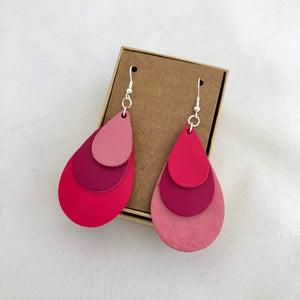 Tri Color Rainbow Earrings in Shades of Pink. This pair of earrings features a mix-matched order of color, so one earring is light pink, magenta, and hot pink, whereas the other is hot pink, magenta and light pink. The earrings are pictured on a brown card in a brown jewelry box, which both come included with the earrings. They are pictured against a white background.
